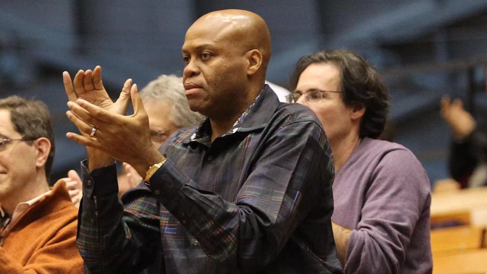 Craig Robinson ’83 stands and applauds in a gymnasium, wearing a dark plaid shirt.
