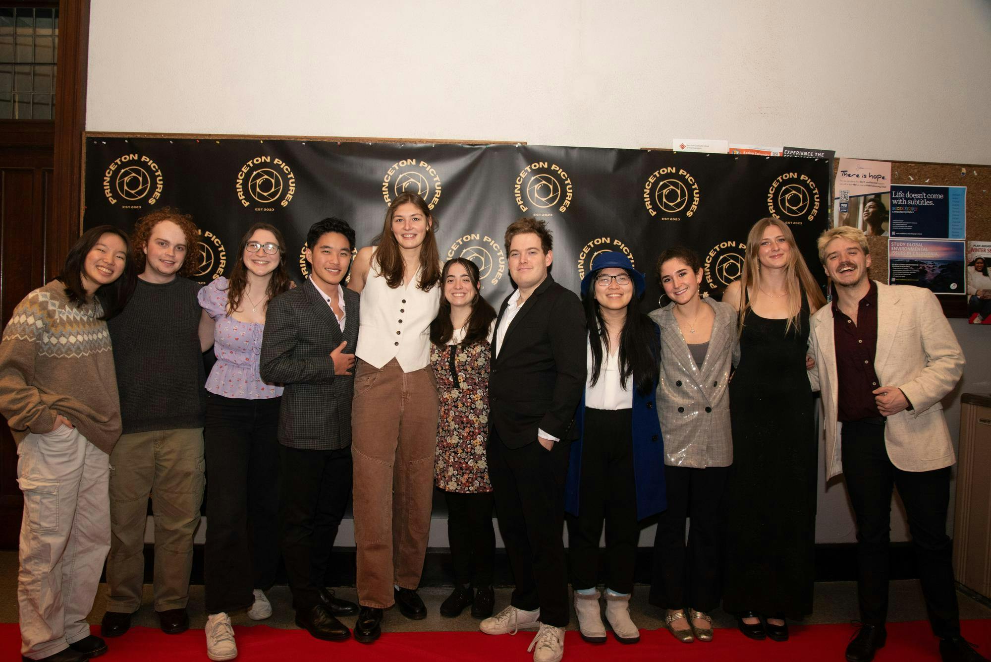 Eleven students pose in front of a black banner with a gold logo pattern. 