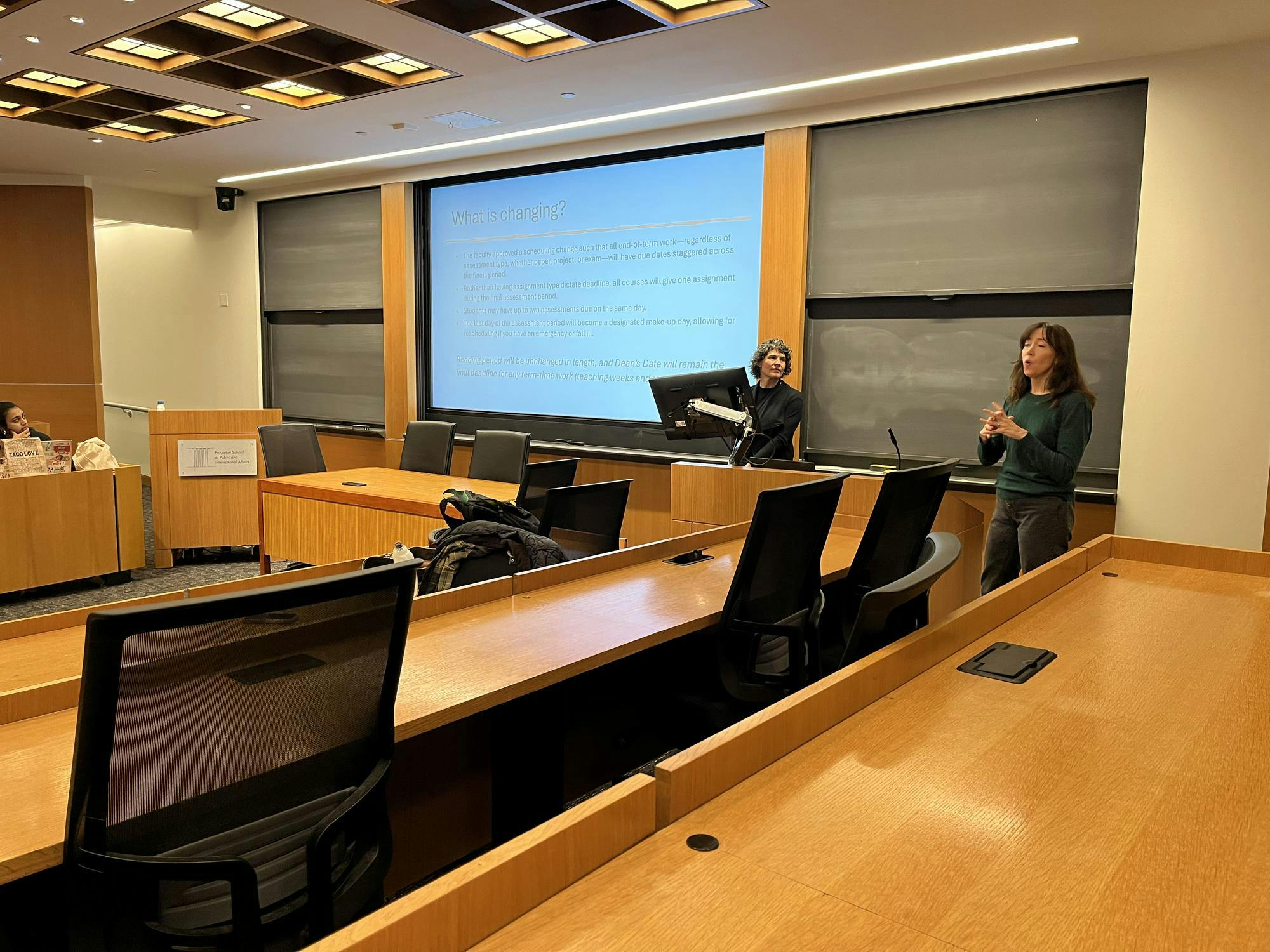 Two women stand in front of a blackboard and smart board combination. The smart board has a presentation which reads "What is changing?" In the foreground, there are wooden desks with black chairs.