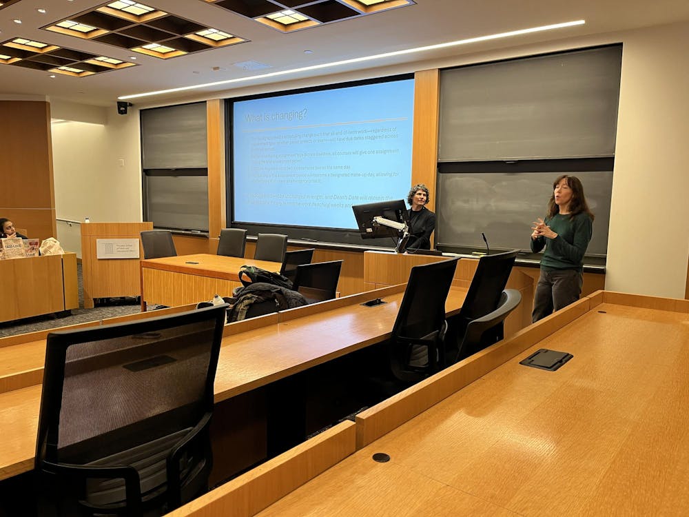 Two women stand in front of a blackboard and smart board combination. The smart board has a presentation which reads "What is changing?" In the foreground, there are wooden desks with black chairs.