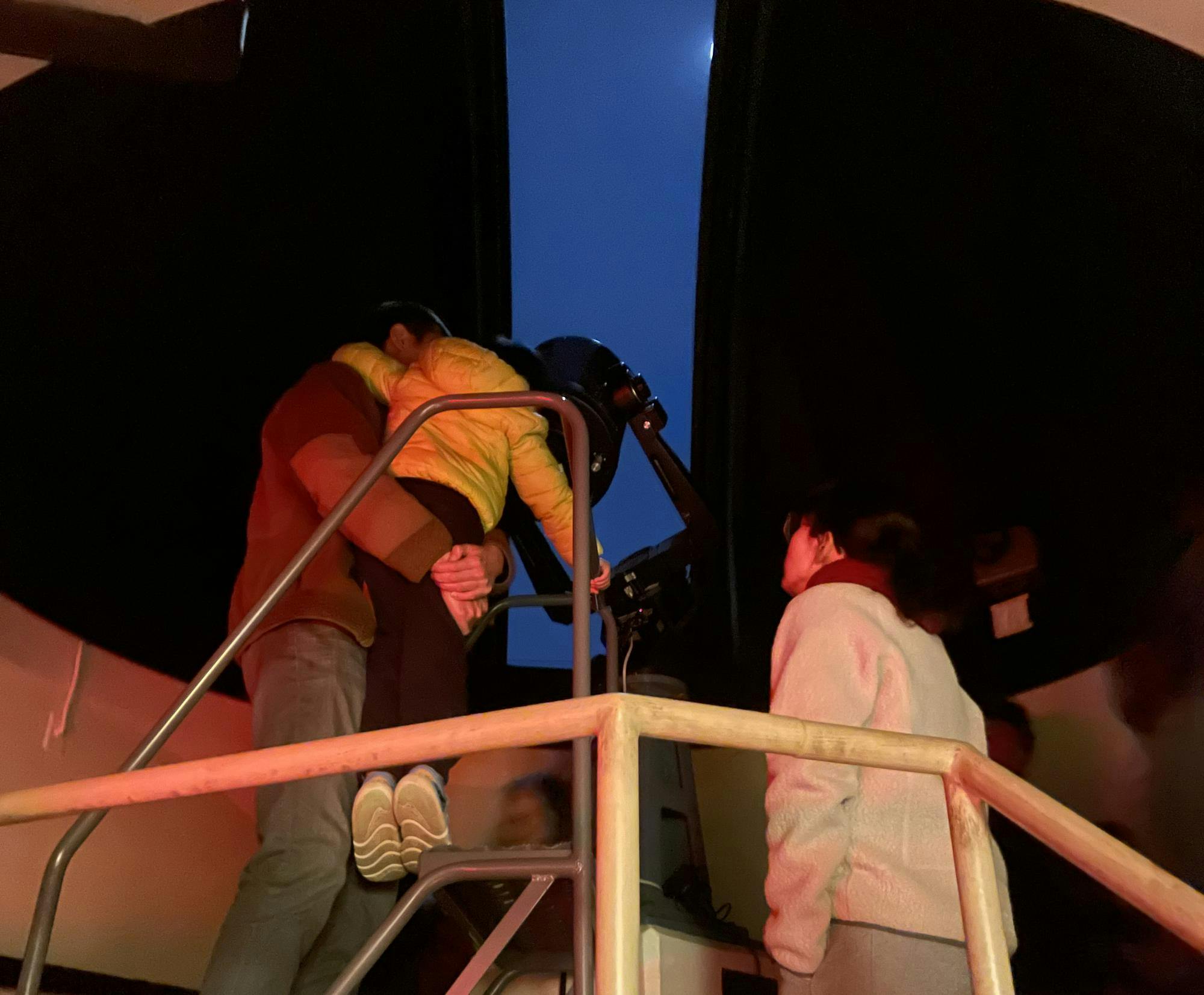 A man wearing a brown jacket holds up a small child wearing a yellow jacket to see through the eye of the telescope. Another person stands behind them, and a sliver of dark blue sky is visible through the observatory ceiling.