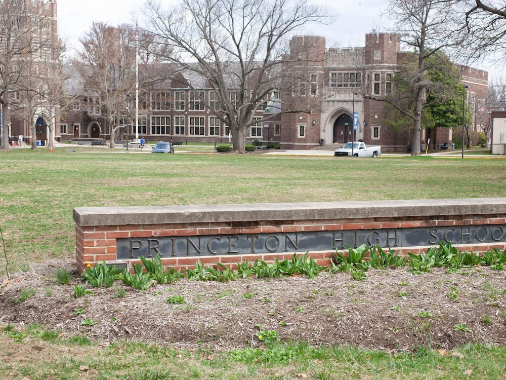 A red-brick building with large windows stands in the background. There is a green field in front of it. In the foreground, there is a brick bench with "Princeton High School" written across it.