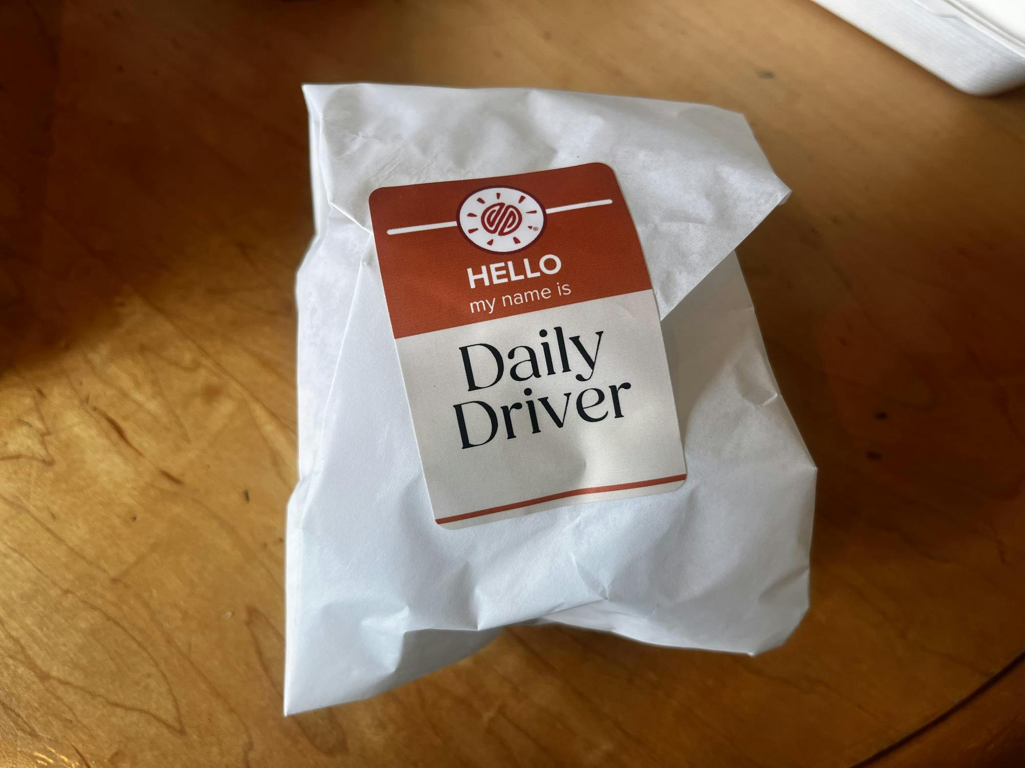 Small World's new burger wrapped in a white package, the red sticker with the Small World Logo reads "HELLO my name is Daily Driver."