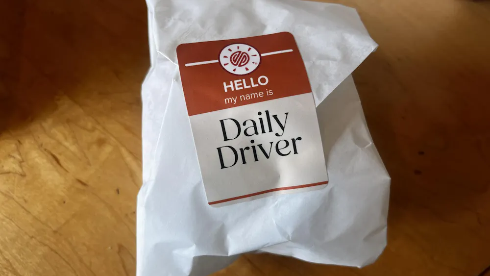Small World's new burger wrapped in a white package, the red sticker with the Small World Logo reads "HELLO my name is Daily Driver."