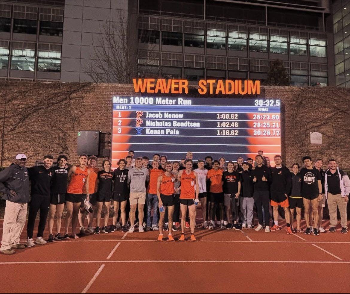 Two Princeton athletes stand arm-in-arm in front of the rest of their team and a scoreboard displaying their times.