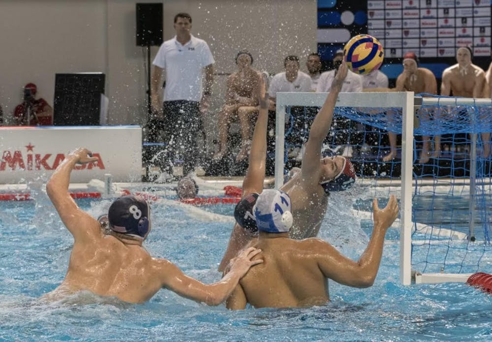 Multiple men are in the water as the goalkeeper with a blue cap extends his left hand to make a save. 