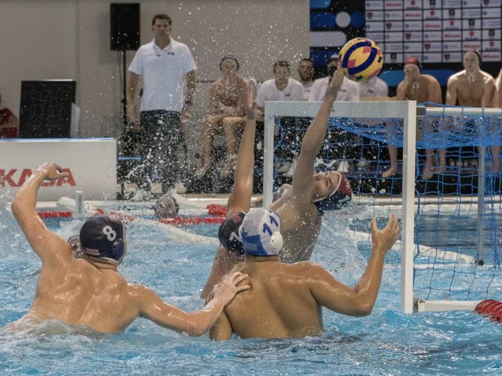 Multiple men are in the water as the goalkeeper with a blue cap extends his left hand to make a save. 