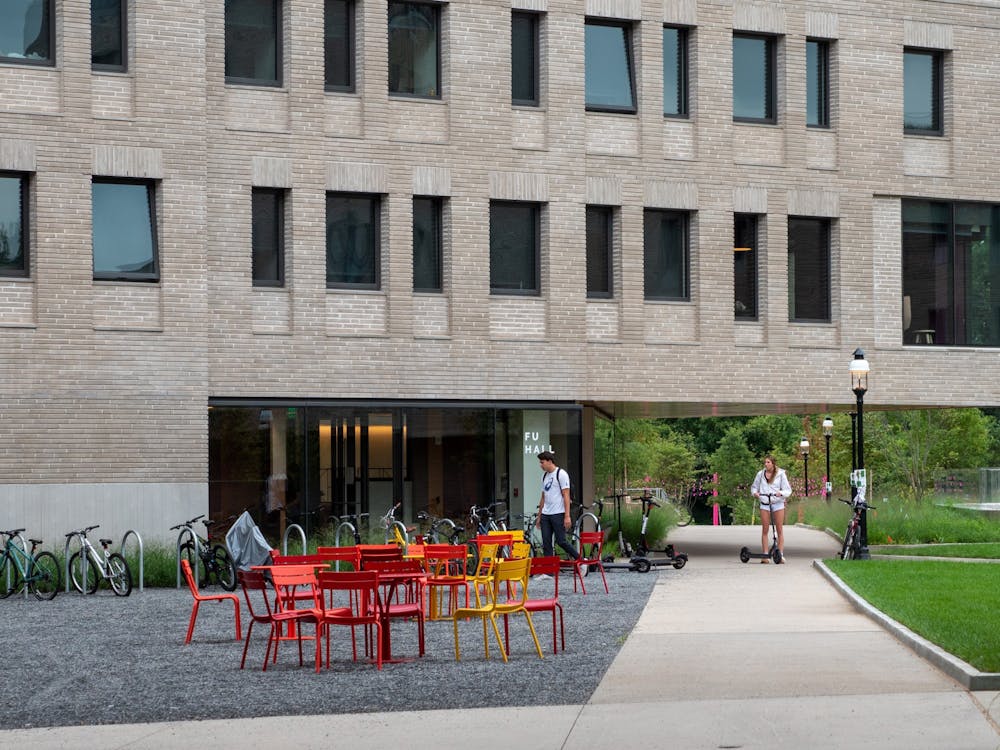 Gray brick modern building with red and white patio furniture above a gravel ground. Two individuals walking with personal electric vehicles along a concrete path.