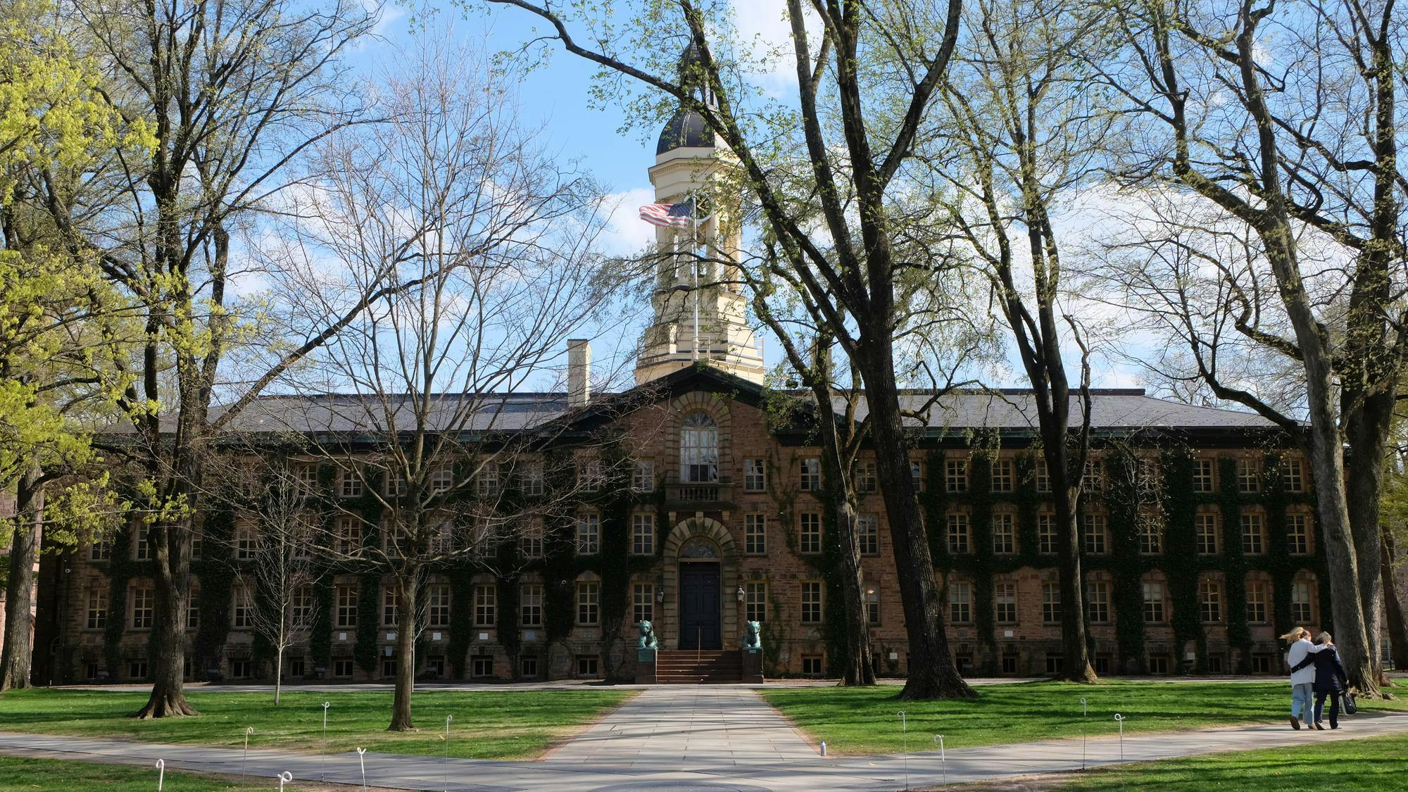 An&#x20;ivy-covered&#x20;brick&#x20;hall&#x20;sits&#x20;at&#x20;the&#x20;end&#x20;of&#x20;a&#x20;paved&#x20;walkway,&#x20;framed&#x20;by&#x20;trees&#x20;and&#x20;grass.