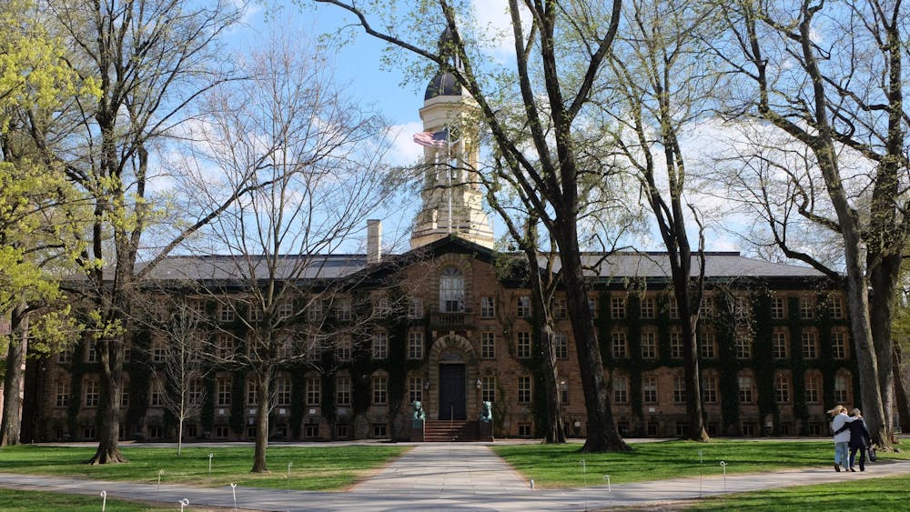 An ivy-covered brick hall sits at the end of a paved walkway, framed by trees and grass.
