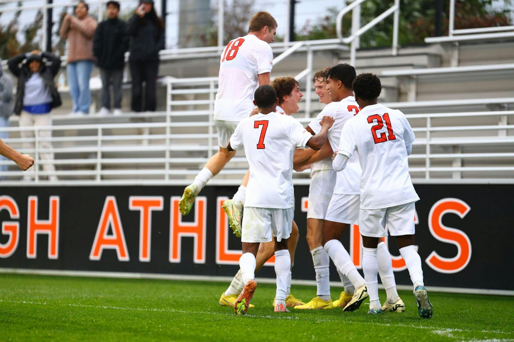 A group of men all wearing white soccer jerseys celebrate on a grass field. 