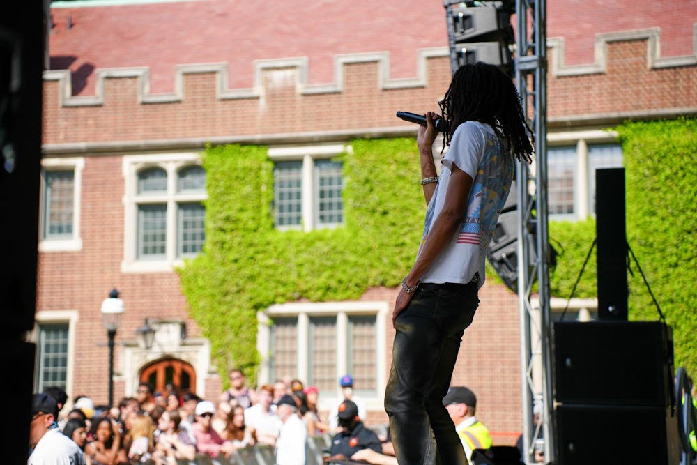 A Black man with dreadlocks performs on stage as a crowd looks on. A brick building with ivy stands in the background.