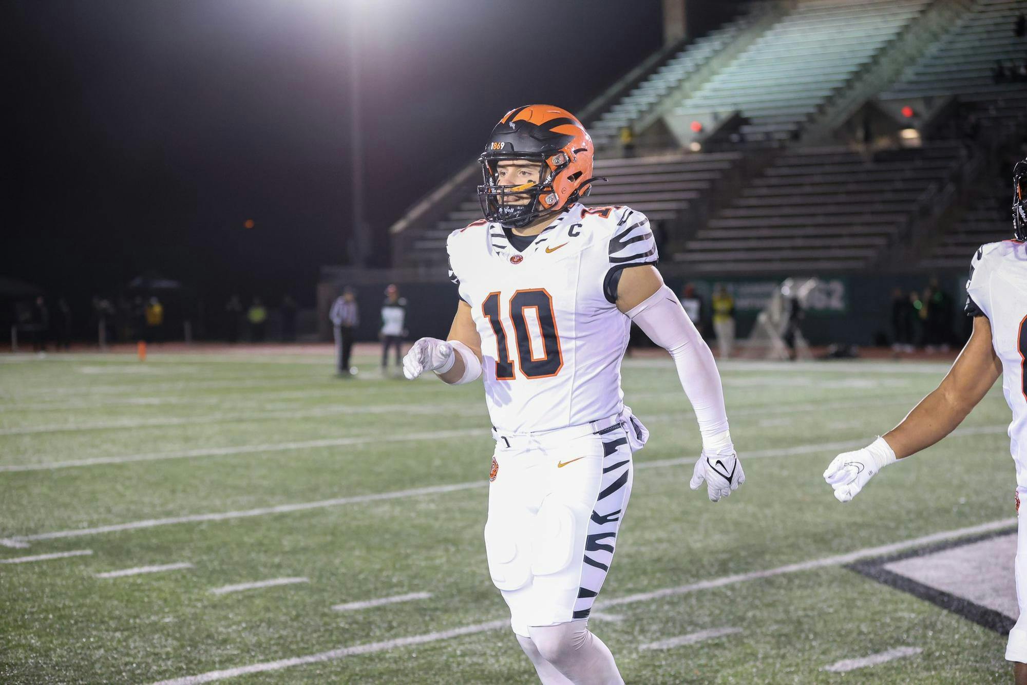A man in a white jersey and orange and black helmet walking off a grass field. 