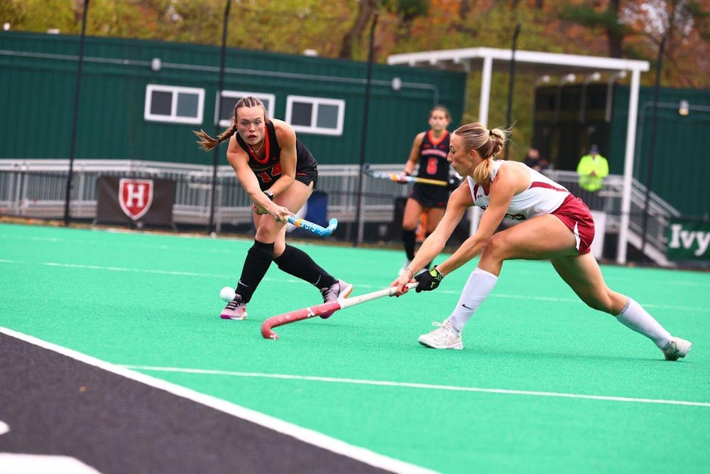 A field hockey player in an orange and black uniform hits the ball past a player on the opposing team.