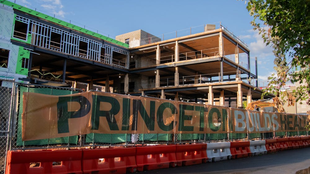 A three story construction site in front of a blue sky with a fence in front with the words on tarp “Princeton Builds Health”