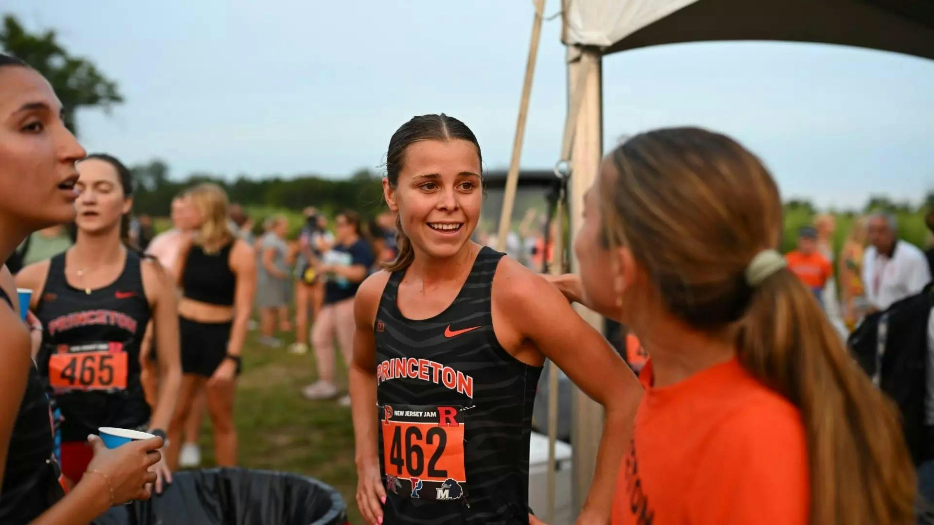 Woman stands in the center of the frame smiling, wearing a black and orange Princeton running tank top. She is looking at another girl wearing an orange shirt.