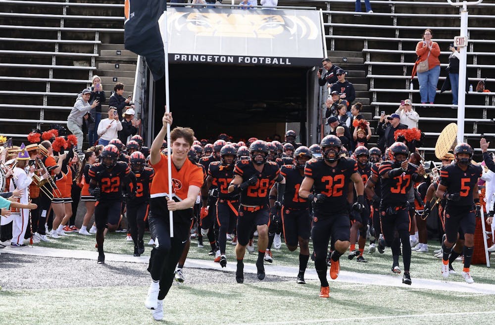 Football players in black jerseys run onto a football field as a cheerleader runs in front of them carrying a flag. 