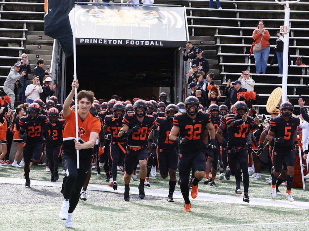 Football players in black jerseys run onto a football field as a cheerleader runs in front of them carrying a flag. 