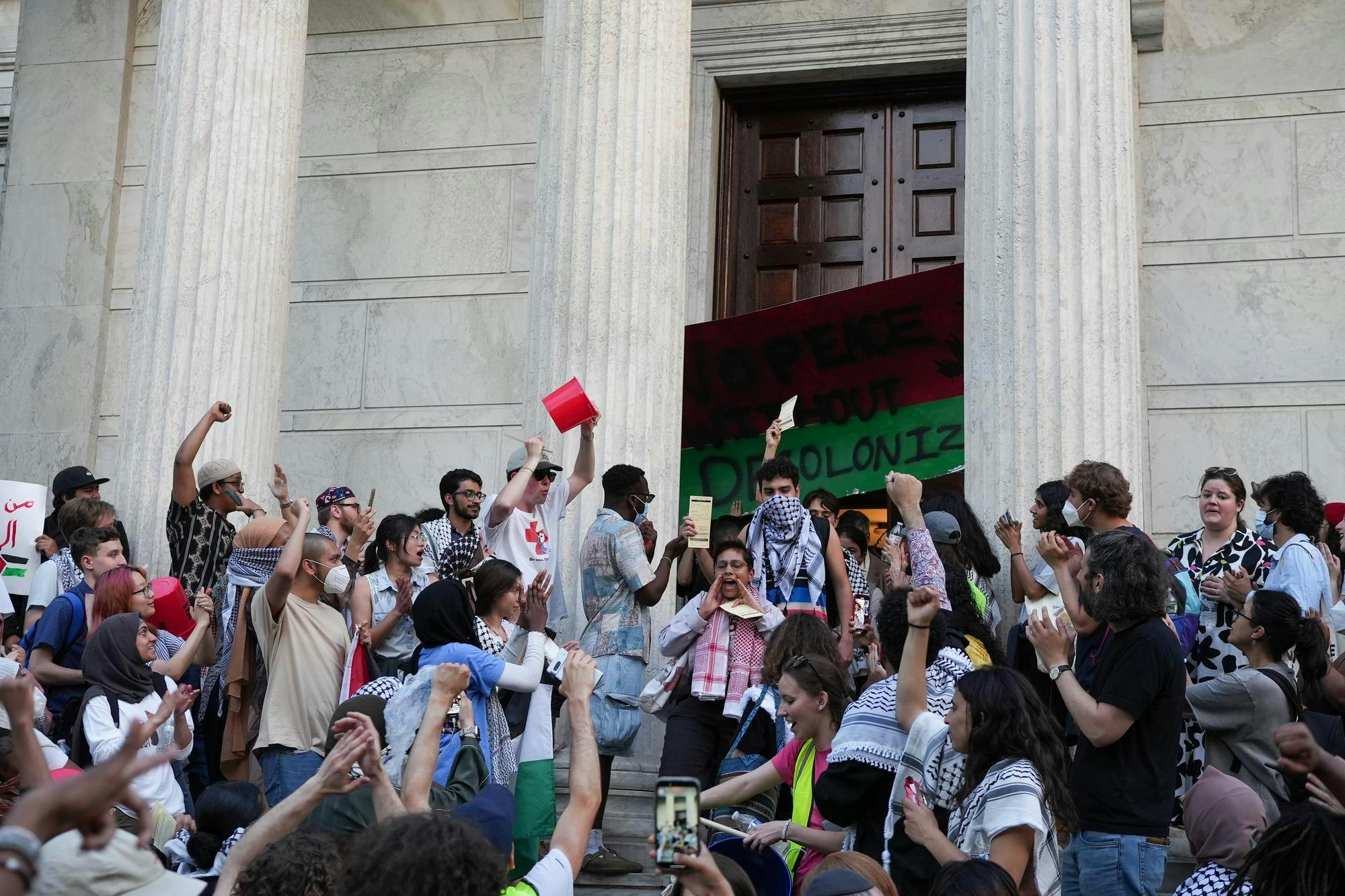 A large group of people surround a white marble building with pillars and shout.