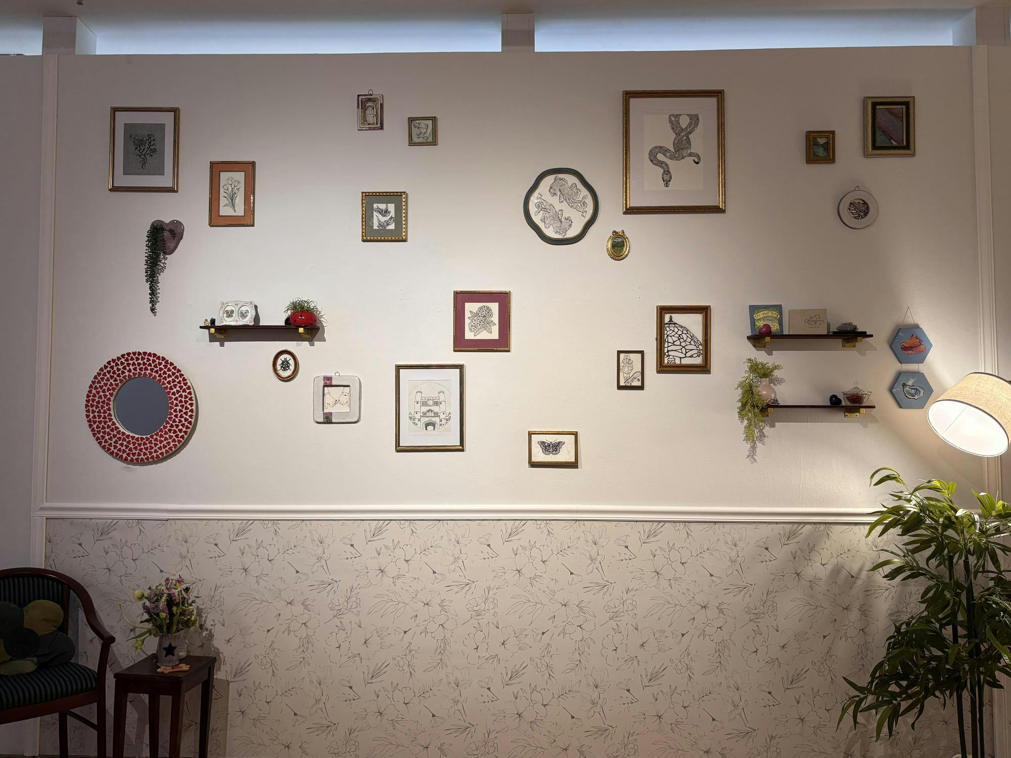 There is a white wall that has floral wallpaper on the bottom. There are a collection of black and white art pieces in multicolored frames. There are also brown hanging shelves with multicolored books and green plants. There is a circular mirror with red dots around the frame.