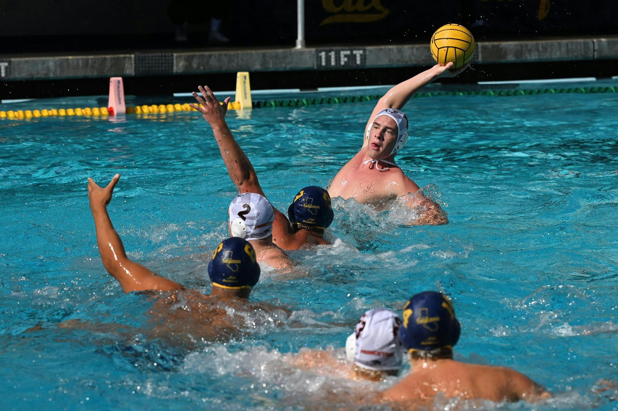 Three men wearing blue and white caps play water polo in a pool filled with aqua colored water.