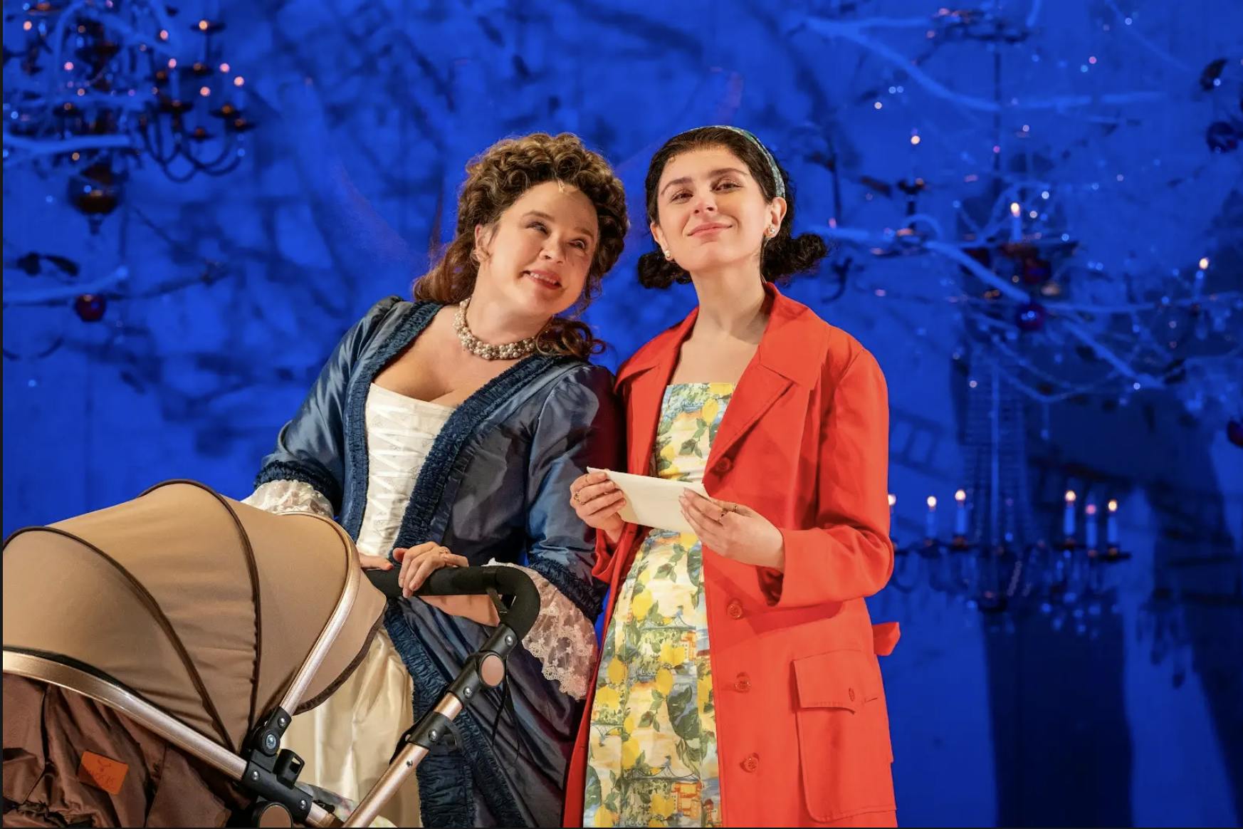 Two women, one in a blue petticoat and white dress and another in a red coat and a yellow dress, smile at each other and look at the camera.