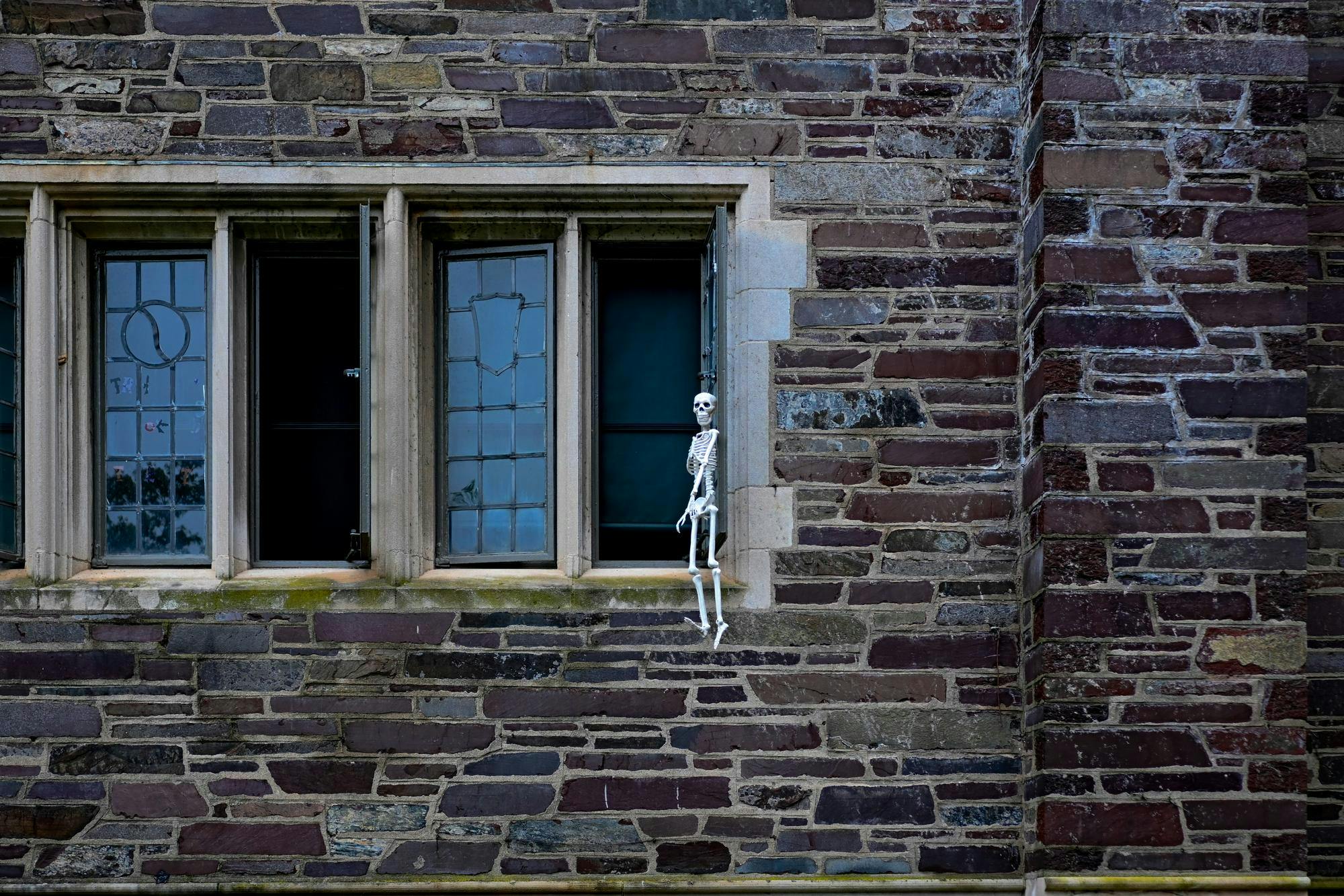 a skeleton hanging outside a window in a grey brick building with four visible windows
