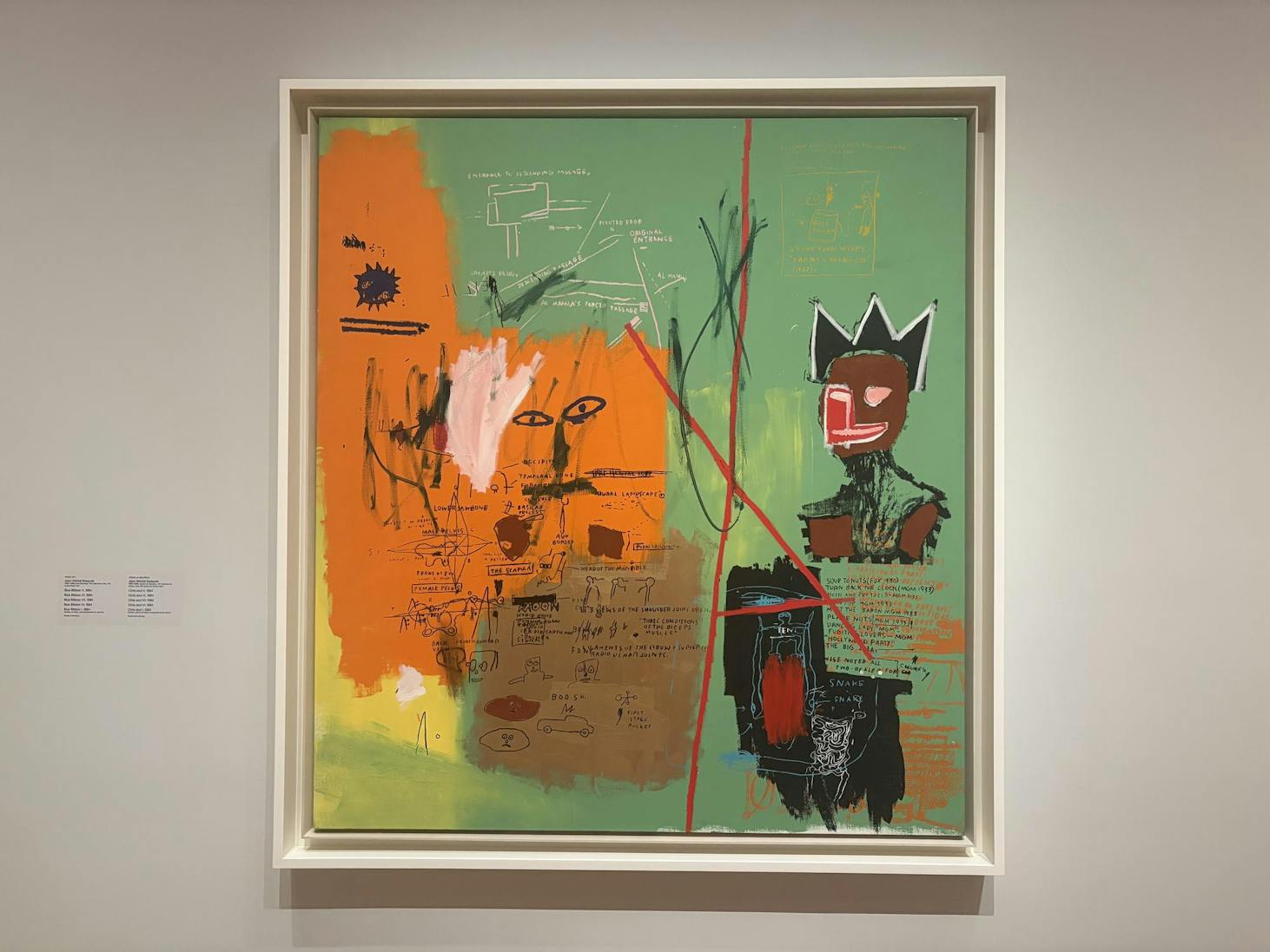 A painting with a green background; orange, brown, yellow, and black patches; and interspersed sketches of anatomical models, phrases, and a figure with a crown is hung against a white wall. 