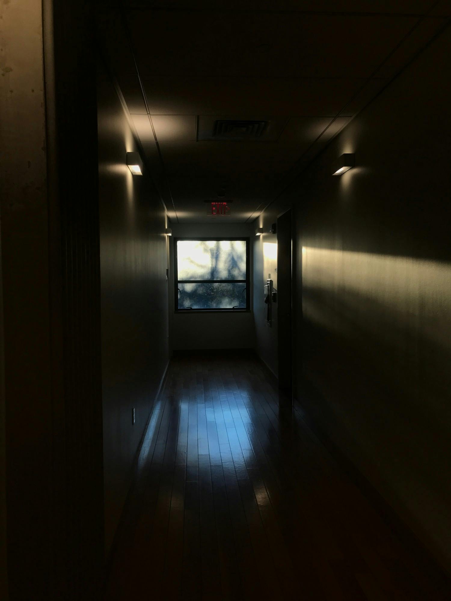 Window viewed from the end of a dark hallway with a door on the far right side and an exit sign hanging above the window.