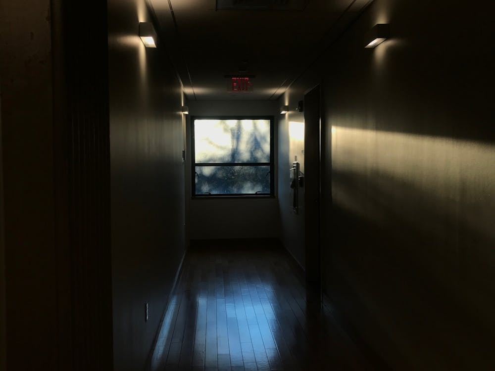 Window viewed from the end of a dark hallway with a door on the far right side and an exit sign hanging above the window.
