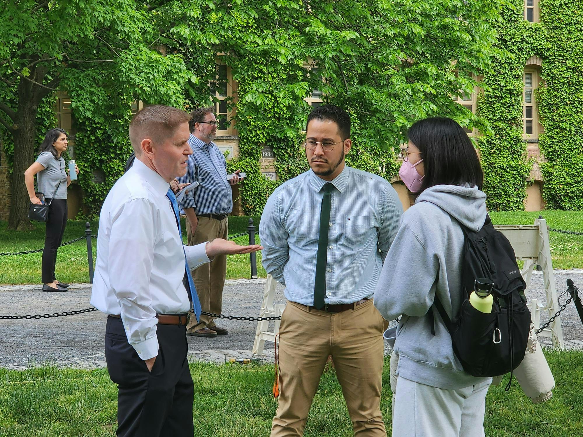 Men in button-down shirts and ties talk to someone in a hoodie and face mask