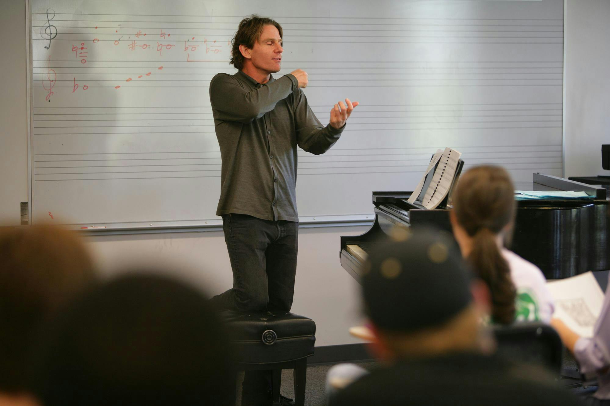 A man teaching a group of students. He stands beside a piano and in front of a whiteboard with music notes drawn on it.