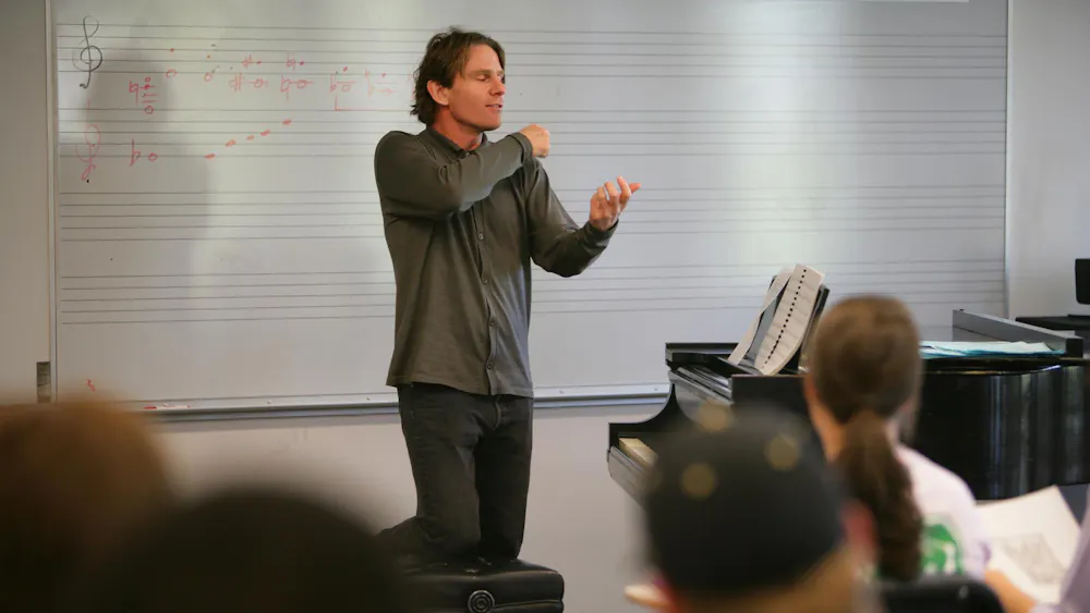 A man teaching a group of students. He stands beside a piano and in front of a whiteboard with music notes drawn on it.