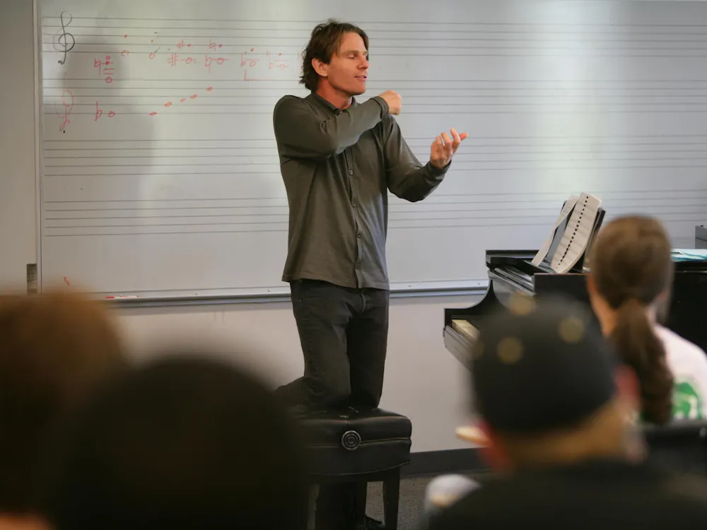 A man teaching a group of students. He stands beside a piano and in front of a whiteboard with music notes drawn on it.