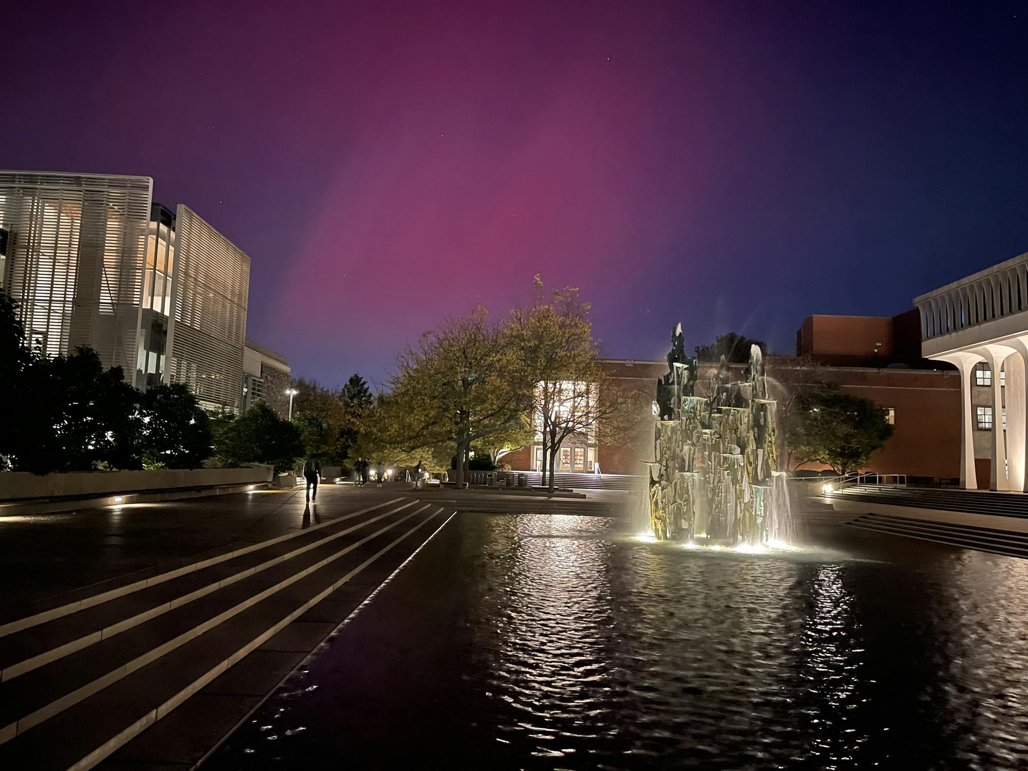 Pink and purple streaks the sky above a fountain. Buildings surround the fountain. 