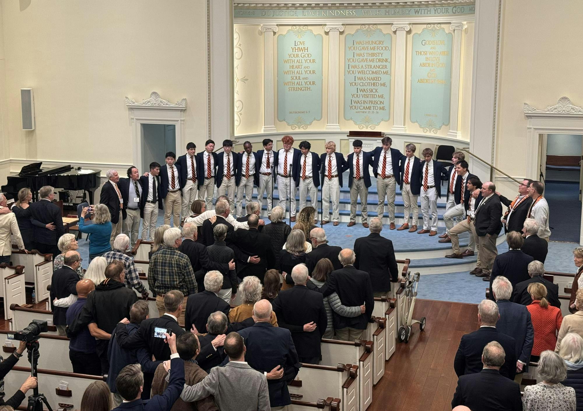 Over a dozen men wearing black suit jackets with orange and black striped ties are standing together on a blue stage in a room with religious text written on the back wall.