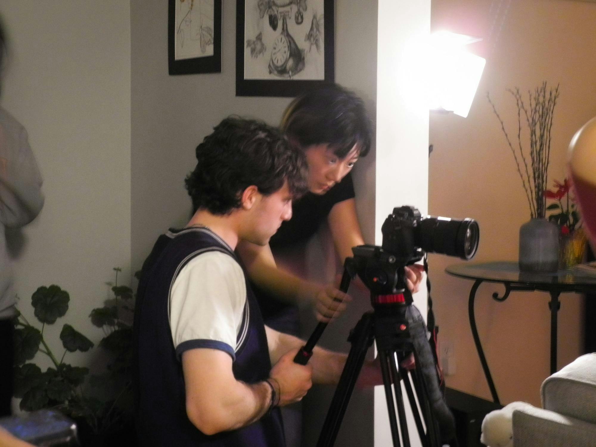 A person in a blue sweater vest and a person in a black shirt are both moving a stationary black camera.