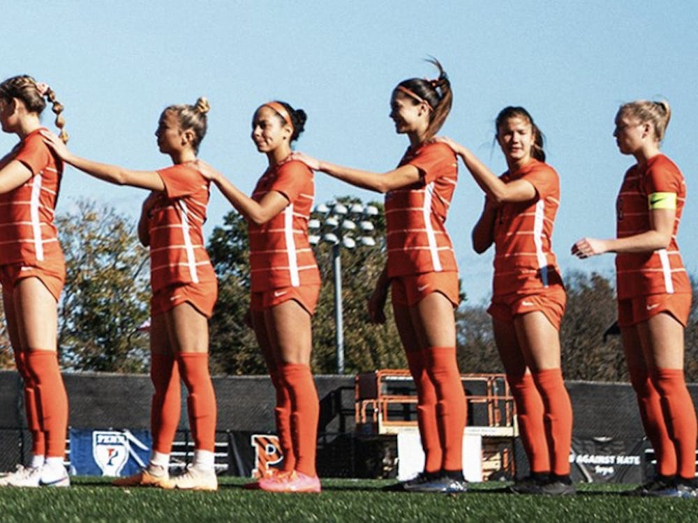women soccer players stand in line with right hand on hard and left hand on left shoulder of player in front of them