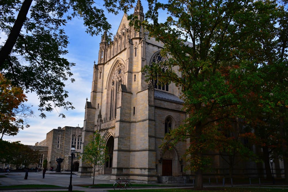 Picture of University chapel with two trees in foreground. Some leaves are orange and yellow, suggesting it’s autumn. A few people are standing in the background.