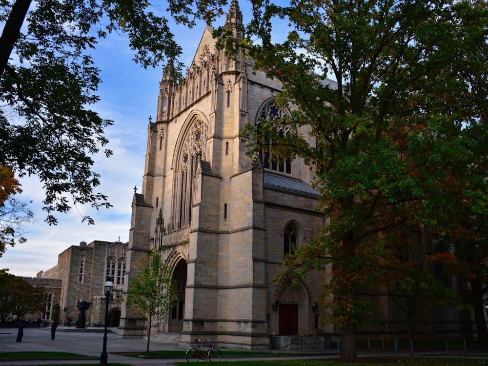 Picture of University chapel with two trees in foreground. Some leaves are orange and yellow, suggesting it’s autumn. A few people are standing in the background.