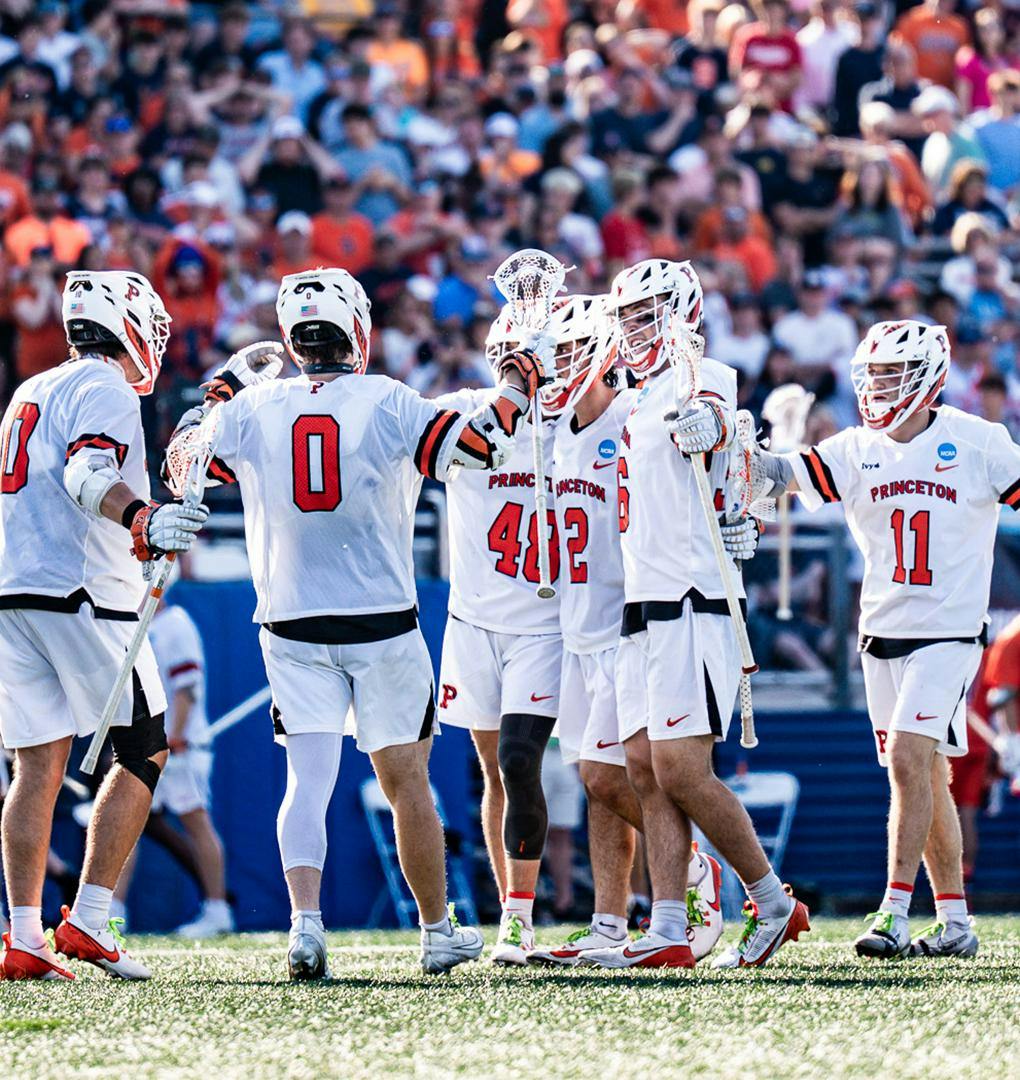 A group of Princeton Men's Lacrosse players in white and orange jerseys circling up together.