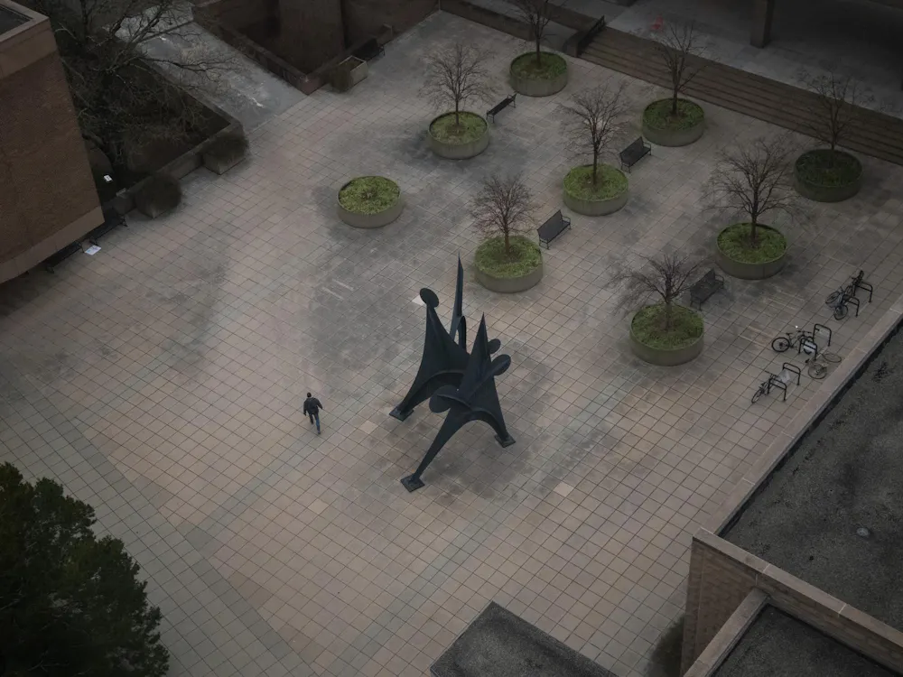 Overhead view of plaza surrounded by buildings; has trees, artistic sculpture, and person walking.