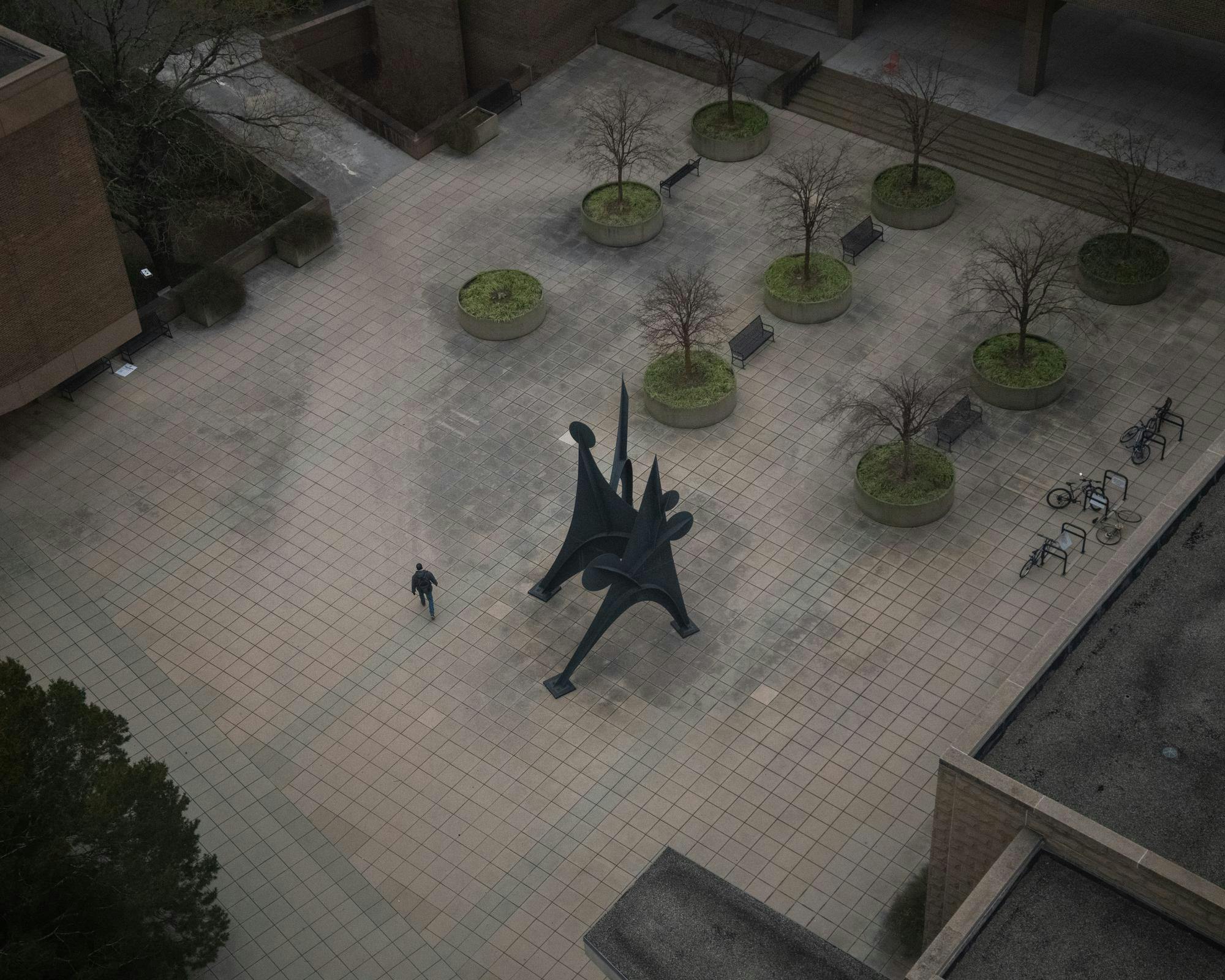 Overhead view of plaza surrounded by buildings; has trees, artistic sculpture, and person walking. 