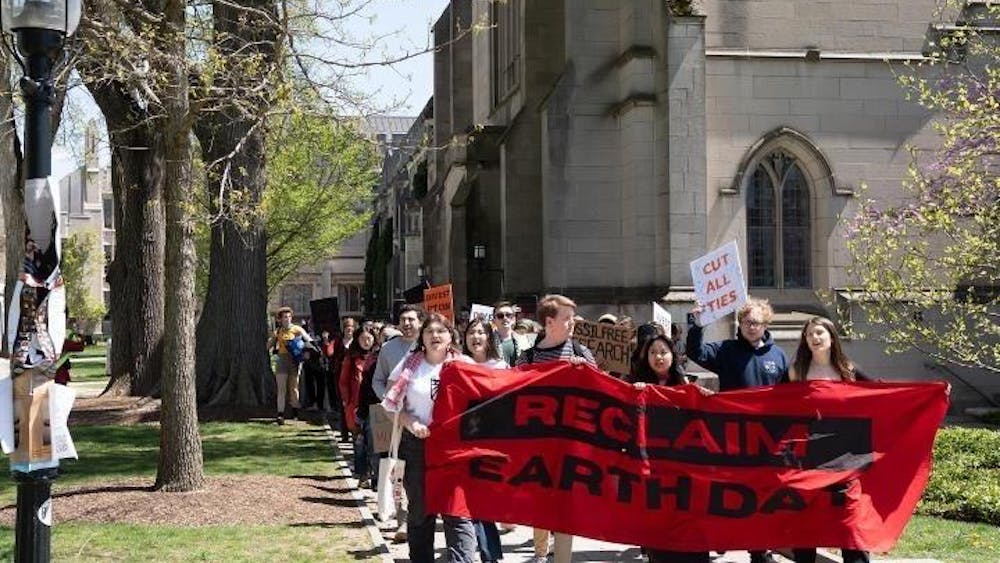 Student protesters in front of stone building holding red banner stating "Reclaim Earth Day" walk in a group. One student protester holds a white sign that says "Cut All Ties." 

