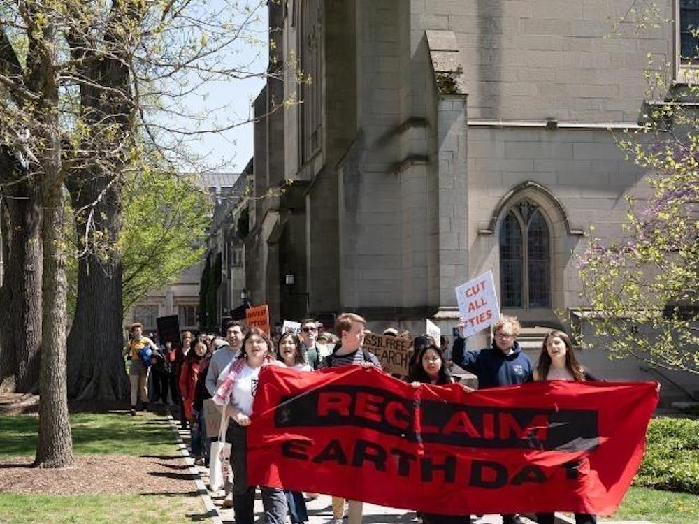 Student protesters in front of stone building holding red banner stating “Reclaim Earth Day” walk in a group. One student protester holds a white sign that says “Cut All Ties.”

