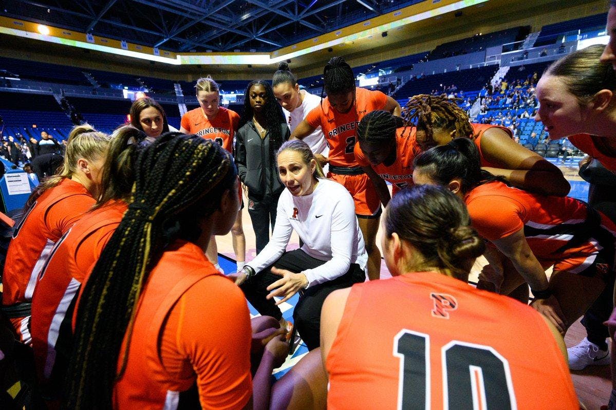 Head coach in white, in center of huddle of players listening to her in orange. 