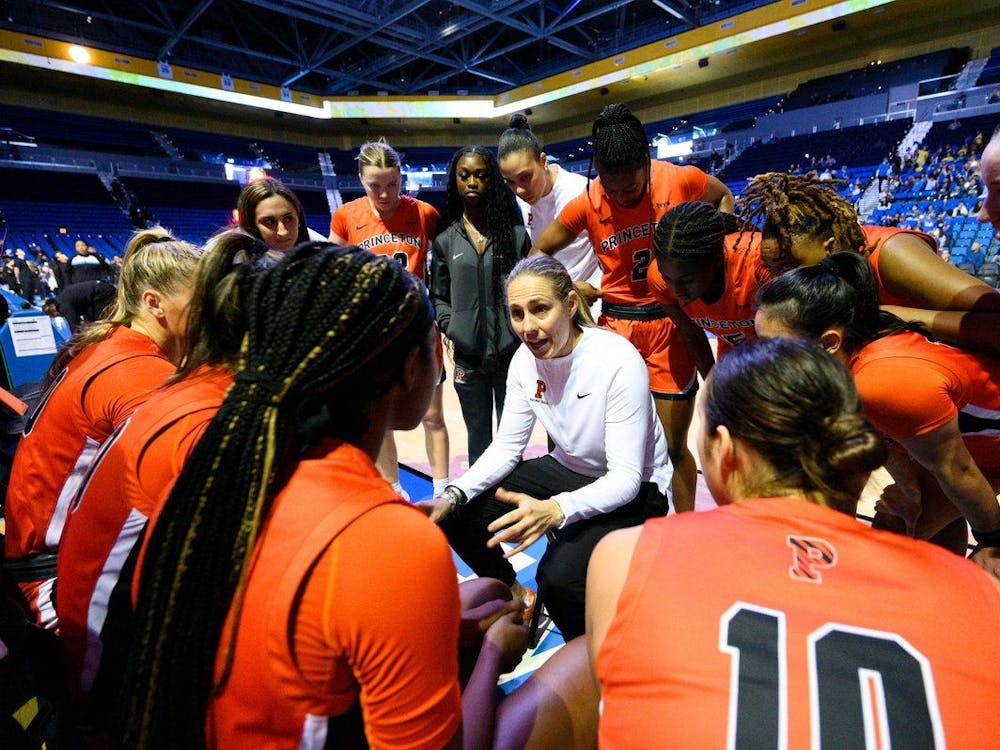 Head coach in white, in center of huddle of players listening to her in orange. 