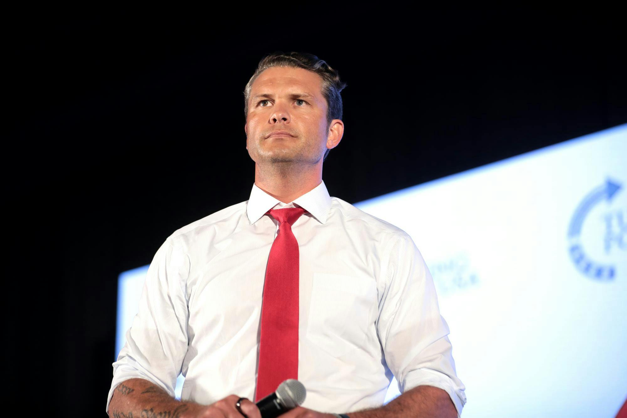 White man wearing a white shirt with red tie on a stage in front of a presentation screen.