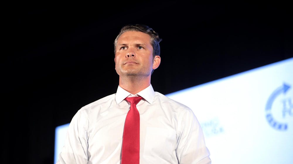 White man wearing a white shirt with red tie on a stage in front of a presentation screen.