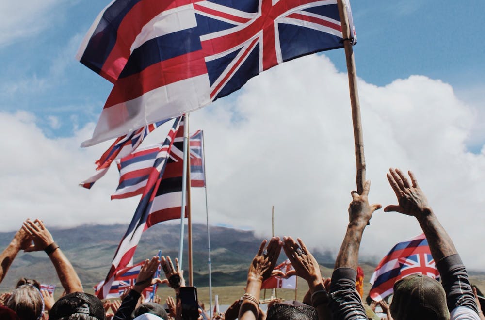 <h5><em><strong>Kia’i</strong></em> <strong>(protectors) in 2019 wave inverted Hawaiian flags to illustrate a nation in distress; their hands, which are in a </strong>‘<strong>triangle shape</strong>’<strong> to indicate a shape of a mountain, highlight their support and love for the Hawaiian people’s sacred</strong> <a href="https://www.honolulumagazine.com/the-sacred-history-of-maunakea/" target="_self"><strong>Mauna Kea</strong></a> <strong>during the Thirty-Meter Telescope (</strong><a href="https://www.hawaiinewsnow.com/2019/07/13/exploring-timeline-leading-up-conflict-mauna-kea/" target="_self"><strong>TMT</strong></a><strong>) protests.&nbsp;</strong></h5>
<h6><strong>Photo courtesy of Monet Bisch</strong></h6>